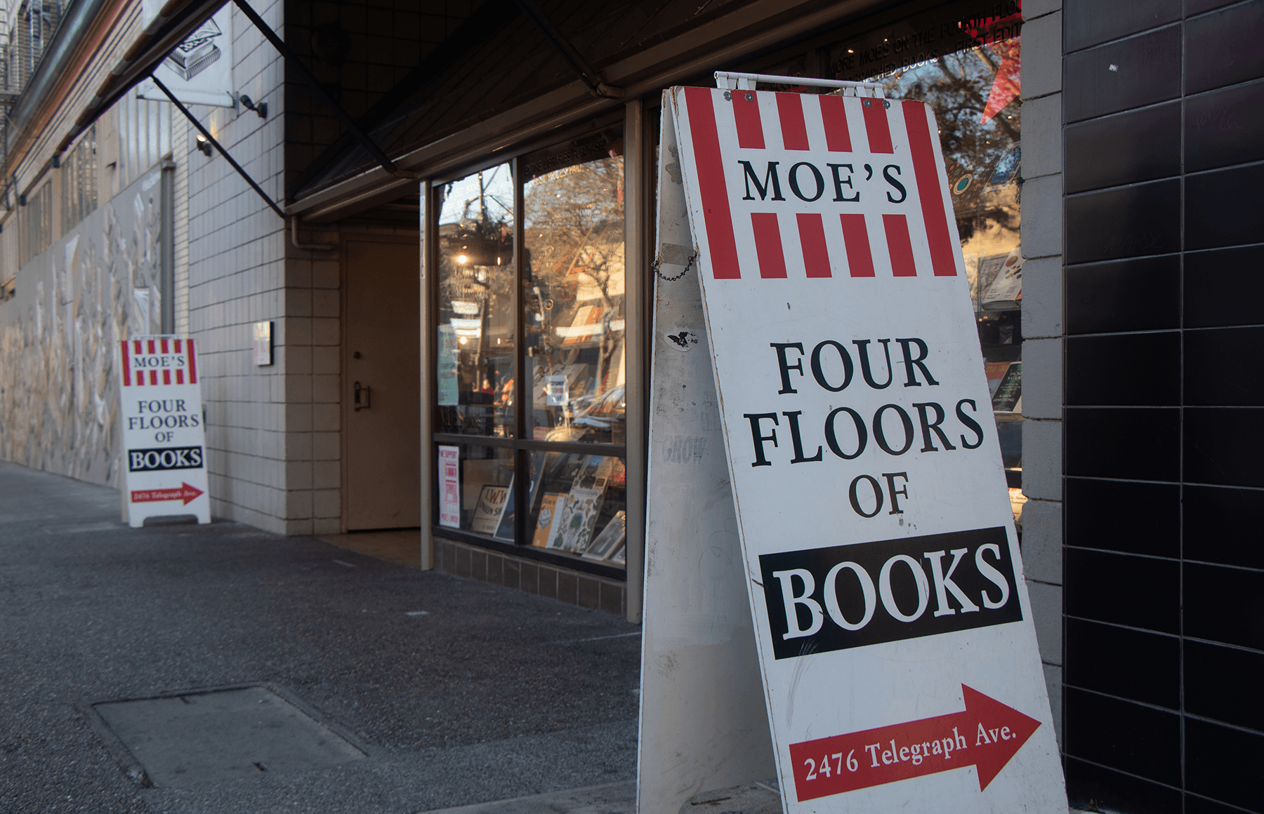 In front of Moe's Book's Telegraph Avenue location, two signs are propped on each side of the storefront. The identical signs have three sections of text. In all capital letters, the first section reads: Moe's. Also in capital letters, the second section reads: four floors of book. In smaller, title case text, the third section reads Moe's address: 2476 Telegraph Ave.